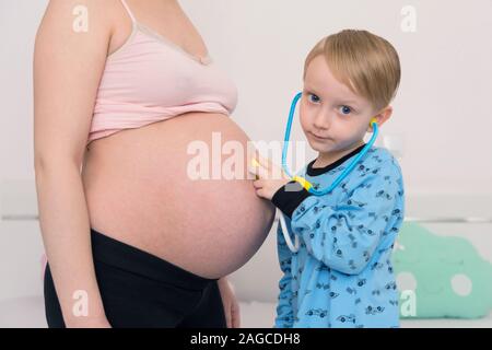 Listening to tummy. Curious handsome dark-eyed boy feeling very interested while listening to pregnant tummy of his pregnant mother. Stock Photo