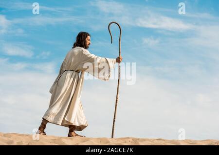 bearded man holding wooden cane and walking in desert against sky Stock Photo