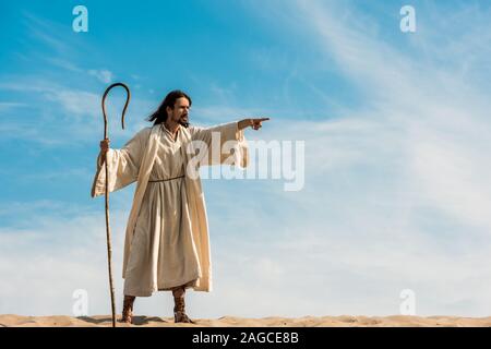 bearded man holding wooden cane and pointing with finger in desert against sky Stock Photo