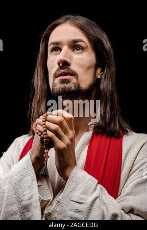 bearded man praying while holding rosary beads isolated on black Stock Photo