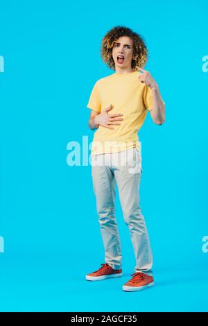 hungry curly teenager pointing with finger at mouth and touching stomach isolated on blue Stock Photo