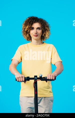 smiling curly teenager riding electric scooter isolated on blue Stock Photo