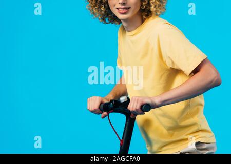 cropped view of smiling curly teenager riding electric scooter isolated on blue Stock Photo