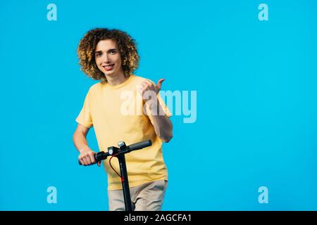 smiling curly teenager riding electric scooter and showing thumb up isolated on blue Stock Photo