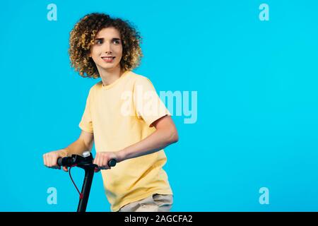 smiling curly teenager riding electric scooter and looking away isolated on blue Stock Photo