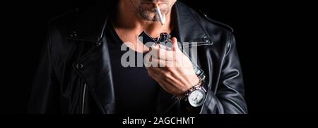 Cropped view of man in leather jacket lighting cigarette with lighter isolated on black, panoramic shot Stock Photo