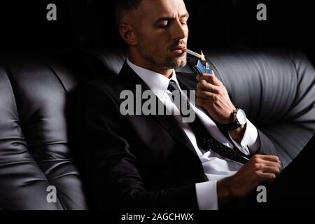 Businessman lighting cigarette while sitting on sofa isolated on black Stock Photo