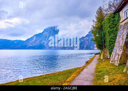 The cozy tiny footpath leads along the bank of Traunsee with medieval building with supports and boasts amazing view on mountain range beyond the lake Stock Photo