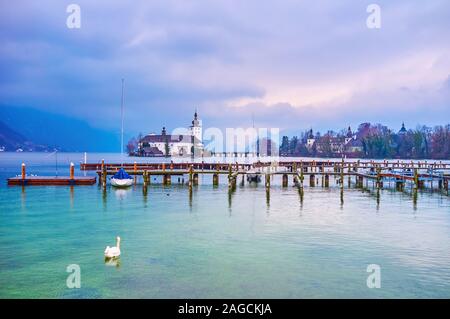 The lonely swan floats at the empty shipyards of Gmunden port on Traunsee lake, the medieval Schloss Ort is seen on the background, Salzkammergut, Aus Stock Photo