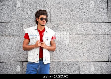 Freestyle. Stylish guy in sleeveless jean jacket sunglasses standing isolated on grey looking aside pensive Stock Photo