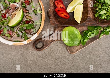 top view of pho in bowl near lime, chili and coriander on wooden cutting board on grey background Stock Photo