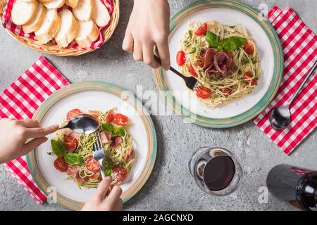 cropped view of women eating Pappardelle with tomatoes, basil and prosciutto near baguette, red wine on grey surface Stock Photo