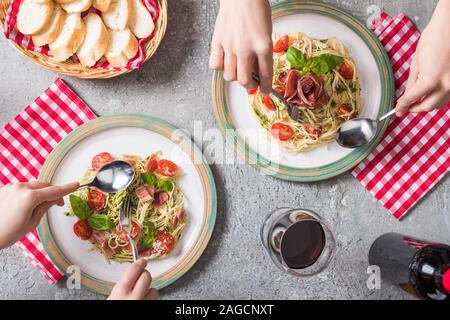 cropped view of women eating Pappardelle with tomatoes, basil and prosciutto near baguette, red wine on grey surface Stock Photo