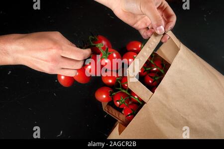 Woman hand taking fresh cherry tomatoes with green vines from brown paper shopping bag over black table, close up detail Stock Photo