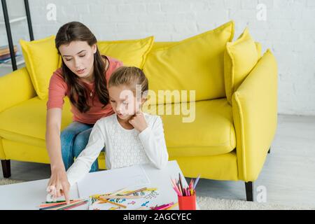 attractive babysitter sitting on yellow sofa near kid and color pencils in living room Stock Photo
