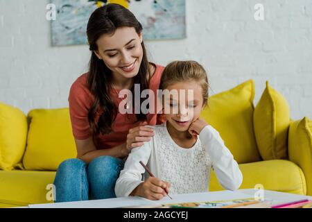 happy babysitter sitting on yellow sofa near kid drawing in living room Stock Photo