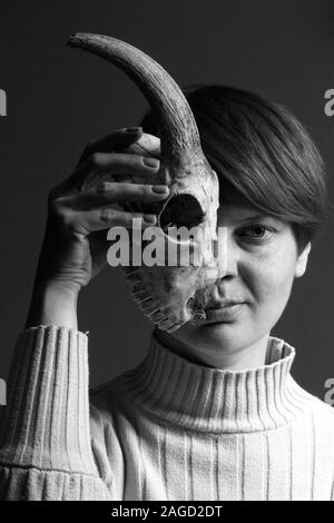 Black and white portrait of a middle-aged redhead woman holding half a goat skull in front of her face, duplicity concept Stock Photo