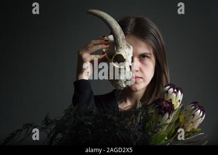 Portrait of a young dark-haired girl holding half a goat skull in front of her face and a bouquet of black proteas and asparagus, duplicity concept Stock Photo