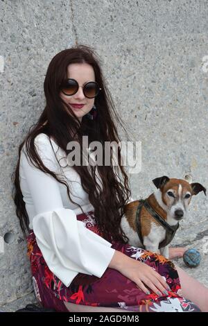A fashionably dressed caucasian lady with long brunette hair and round sunglasses (shades) sits by a concrete wall with her white and tan Jack Russell Stock Photo