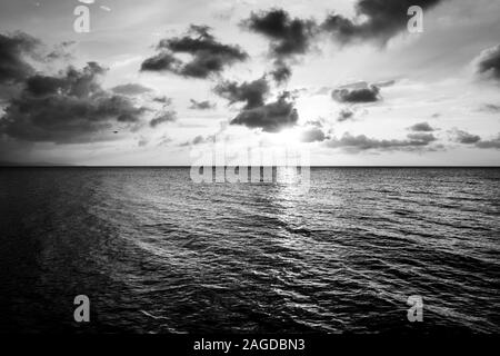 Mesmerizing greyscale shot of the sun setting behind the clouds over the calm ocean Stock Photo