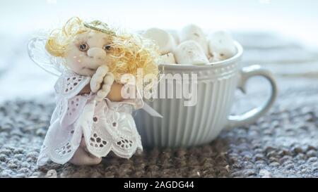 angel in white lace dress with hands folded in prayer. homemade sock toy with fluffy red hair in background of window. cup of marshmallows on knitted Stock Photo