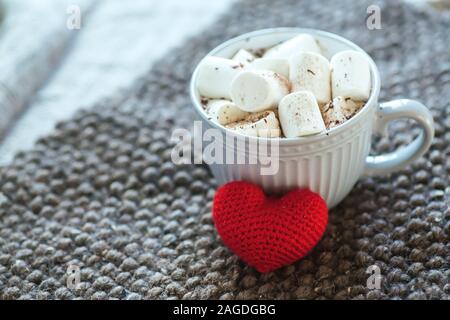 Marshmallow in grey cup of hot cocoa. warm cozy background. Hand-knitted heart from red threads. Valentines day concept, mug cup of coffee Stock Photo