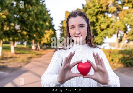 teenager girl holds red toy heart in her hands, helps gift donation, donate support, care love, emotions tenderness comfort. Smiling summer park, free Stock Photo