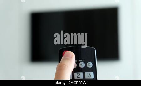 Thumb pressing on the power button of a television remote control, pointing towards a tv in the blurred background. Stock Photo