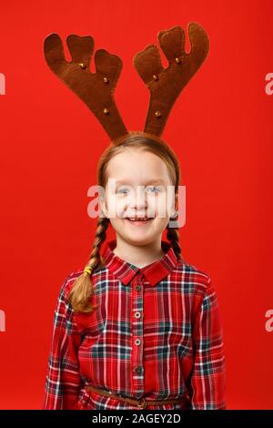 Happy funny little girl in christmas reindeer costume on color red background. Stock Photo