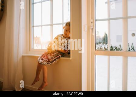 young girl looking sad sitting on a window sill at home at sunset Stock Photo