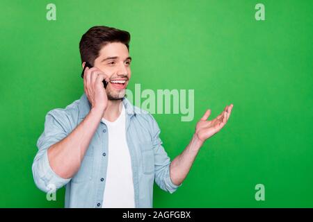Photo of handsome guy holding telephone hand near ear listen best friend humorous story overjoyed good connection wear denim shirt isolated green Stock Photo