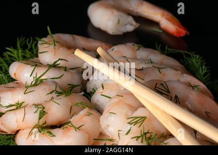 Close up view of freshly cooked peeled prawns or shrimps and chopsticks on a bed of dill on black blackground. Stock Photo