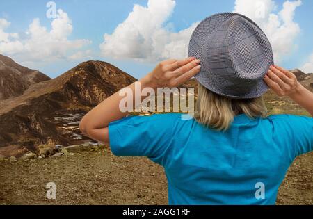 Girl in hat looks at mountain terrain and thick clouds and blue sky from a cliff. White middle aged woman stays back to us and touches her hat’s flaps Stock Photo