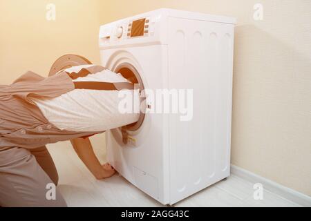 skilled employee in grey uniform gets into repairing broken washing machine on wooden floor at beige wall backside view Stock Photo