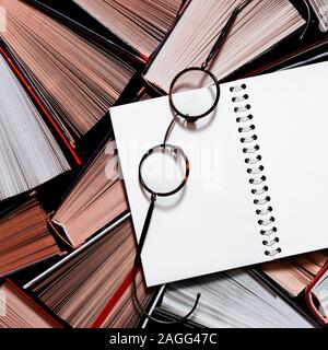 Many multicolored thick open books stand on a dark background. On the books are old round glasses and an open notebook Stock Photo
