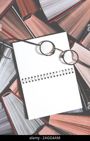 Many multicolored thick open books stand on a dark background. On the books are old round glasses and an open notebook Stock Photo
