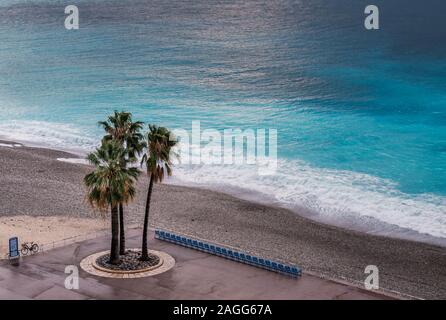 Three palm trees stand next to an empty row of chairs on the sea front on the Promenade des Anglais in Nice against the turquoise Mediterranean sea Stock Photo