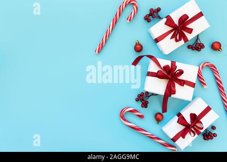 Gift boxes wrapped white paper and red ribbon decorated berries, balls and candy canes on blue background. Top view. Flat lay.  Christmas and New Year Stock Photo