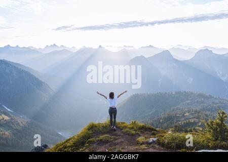 Female hiker raising arms against sun on peak, Winchester Mountain, North Cascades, Washington, USA Stock Photo