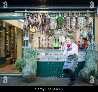 A male butcher standing outside his shop window with pheasants and rabbits hanging up for display Stock Photo