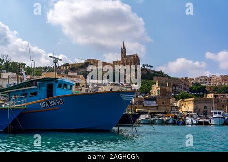 Lourdes Chapel on the promontory, with nose of blue boat in the foreground in Mgarr harbour. Stock Photo