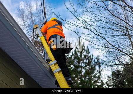 A domestic fiber optic installation is in progress as a workman is seen in full personal protective equipment, PPE, working at height from a ladder Stock Photo