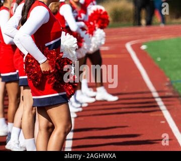 High school cheerleaders standing on the track holding red and white pom poms while watching the football game. Stock Photo