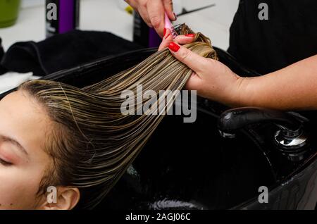 Hair stylist applying hair mask and combing clients wet hair with brush in a black sink. Stock Photo