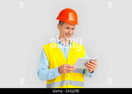 Construction. Mature man in hardhat and uniform standing isolated on white watching video on digital tablet smiling cheerful Stock Photo