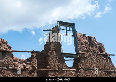 Brick wall with adobe clay plaster and broken wooden window frame remained of ruined old rural country house on cloudy blue sky background Stock Photo