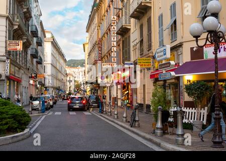 French citizens and tourists walk through a colorful area of shops, hotels and cafes near a police station as lights come on at dusk in Nice, France Stock Photo