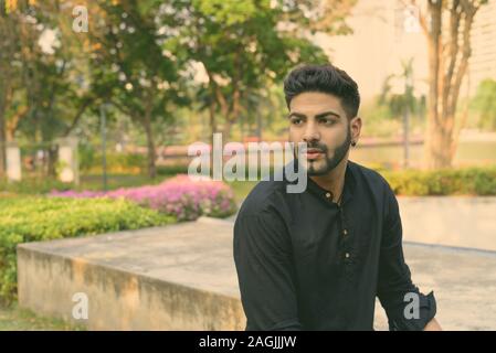 Young handsome Indian man thinking while sitting on concrete floor at the park Stock Photo