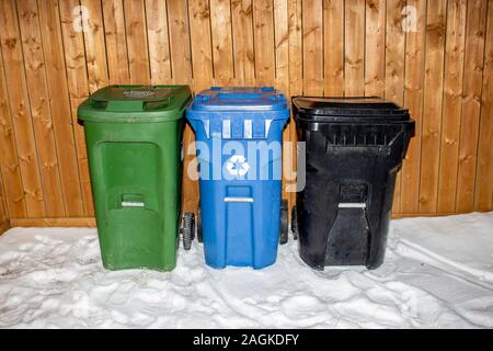 A Green cart for composting, Blue cart for recycling and a black cart for garbage sitting on a back alley during winter Stock Photo