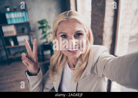 Close up photo of positive cheerful blonde hair middle aged businesswoman leader make selfie greet her clients partners investors make v-sign in Stock Photo